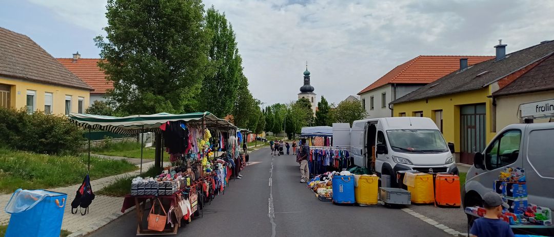 Eine Straße mit Straßenständen und einem Kirchturm im Hintergrund. Kleidung und Handtaschen sind auf den Ständen ausgestellt. Ein Lieferwagen ist in der Nähe geparkt.