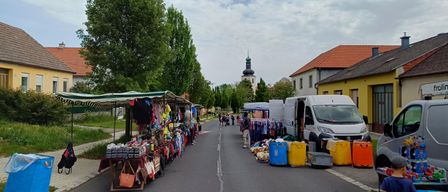A street with sidewalk stalls and a church tower in the background. Clothes and handbags are displayed on the stalls. A van is parked nearby.