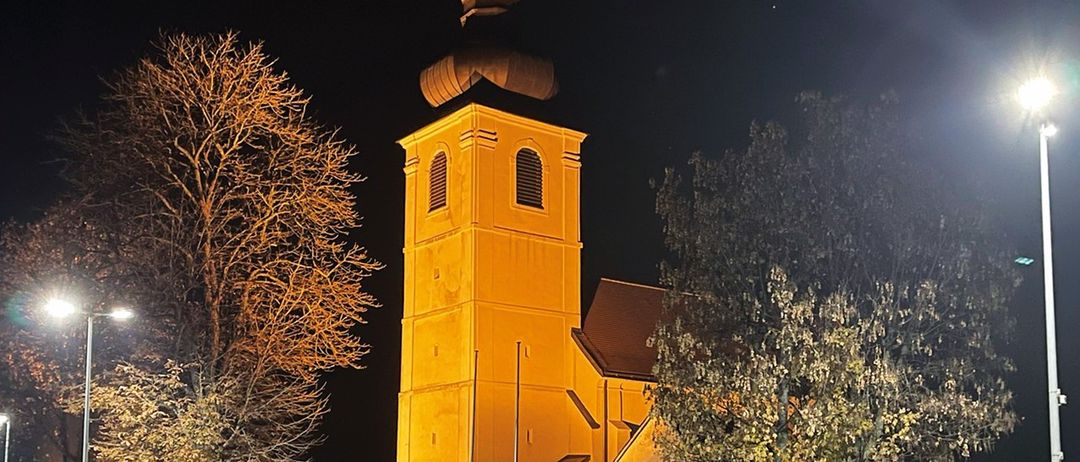 Ein hoher gelber Kirchturm mit einem Kreuz oben, beleuchtet in der Nacht. Der Turm ist von Bäumen und einer Mauer umgeben, mit Bänken und Straßenlaternen in der Nähe.