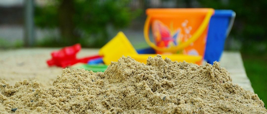 Ein Sandkasten mit einem großen Haufen Sand davor, mit einem farbenfrohen Eimer, Schaufel und anderen Spielzeugen in der Nähe.