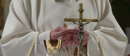 Ein Priester hält ein Kreuz und einen Kelch, beide golden, während einer religiösen Zeremonie. Die Hände des Priesters sind sichtbar, er hält das Kreuz.