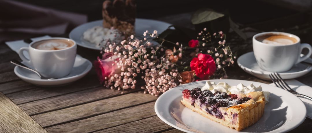 Ein Stück Brombeertarte auf einem weißen Teller, begleitet von Kaffeetassen, Dessert und Blumen auf einem Holztisch.