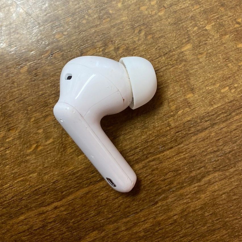 Ein weißer kabelloser Ohrhörer liegt auf einer Holzoberfläche und zeigt sein schlankes Design. Der Ohrhörer hat ein kleines Mikrofon und einen Ladeanschluss.