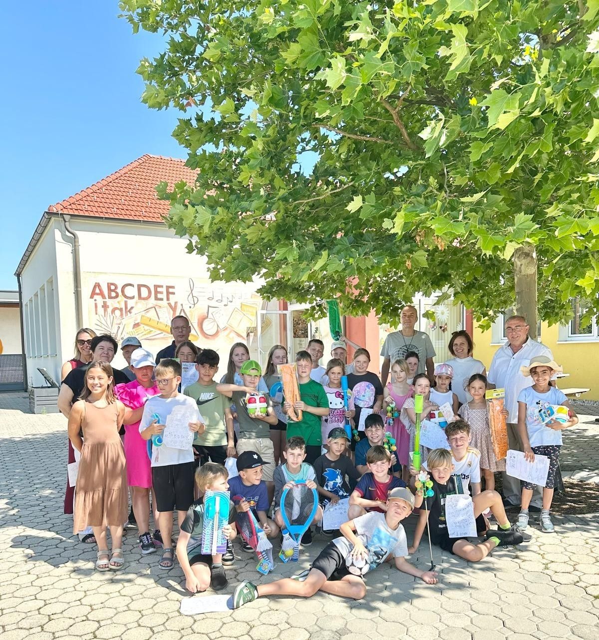 Eine Gruppe von Kindern und Erwachsenen posiert für ein Foto vor einem Gebäude. Das Gebäude hat ein Banner mit ABCDEF itak und Musiknoten. Die Kinder halten Schläger und Papiere in den Händen.