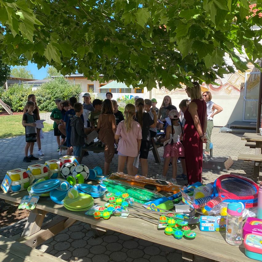 Eine Gruppe von Kindern und Erwachsenen versammelt sich um einen Picknicktisch voller verschiedener Spielzeuge und Spiele unter einem großen Baum.