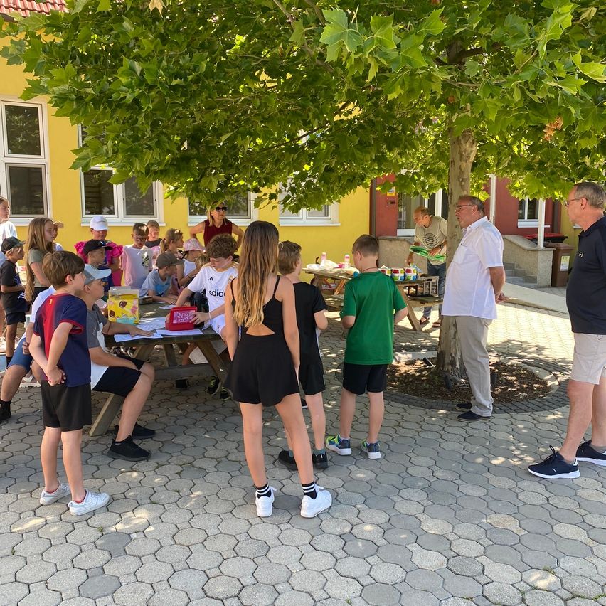 Eine Gruppe von Kindern und Erwachsenen versammelt sich unter einem Baum. Einige sitzen um einen Picknicktisch mit Essen herum, während andere stehen. In der Nähe befindet sich ein gelbes Gebäude mit Fenstern und einer Tür.