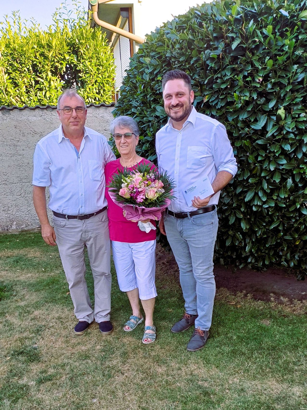 Drei Personen stehen auf einem Rasen. Eine Frau hält einen Blumenstrauß und ein Mann ein Zertifikat. Dahinter ist eine Wand mit grünen Büschen.