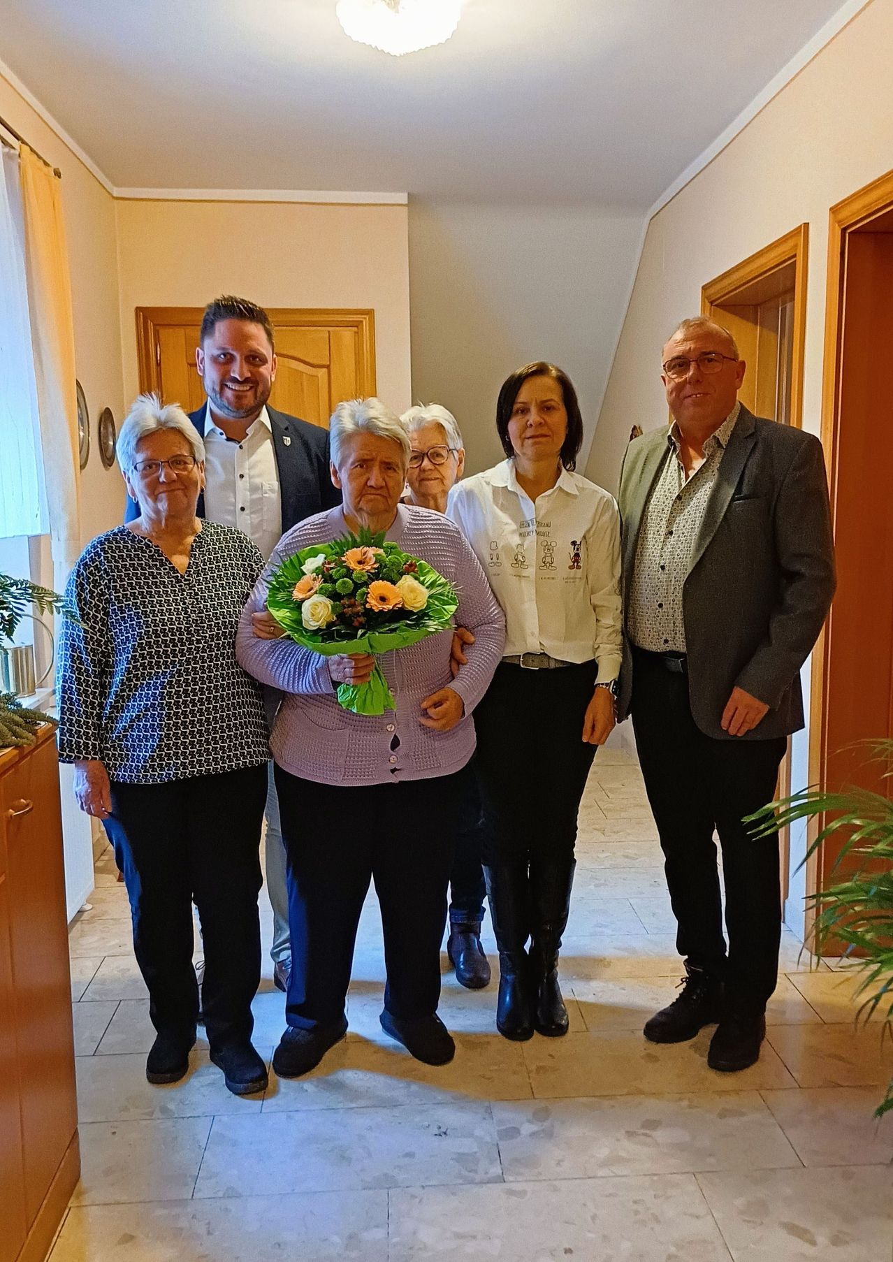 Eine Gruppe von Menschen steht in einem Flur. Eine ältere Frau hält einen Blumenstrauß. Hinter ihr stehen drei Frauen und zwei Männer. Einer der Männer trägt eine Brille und einen Anzug.