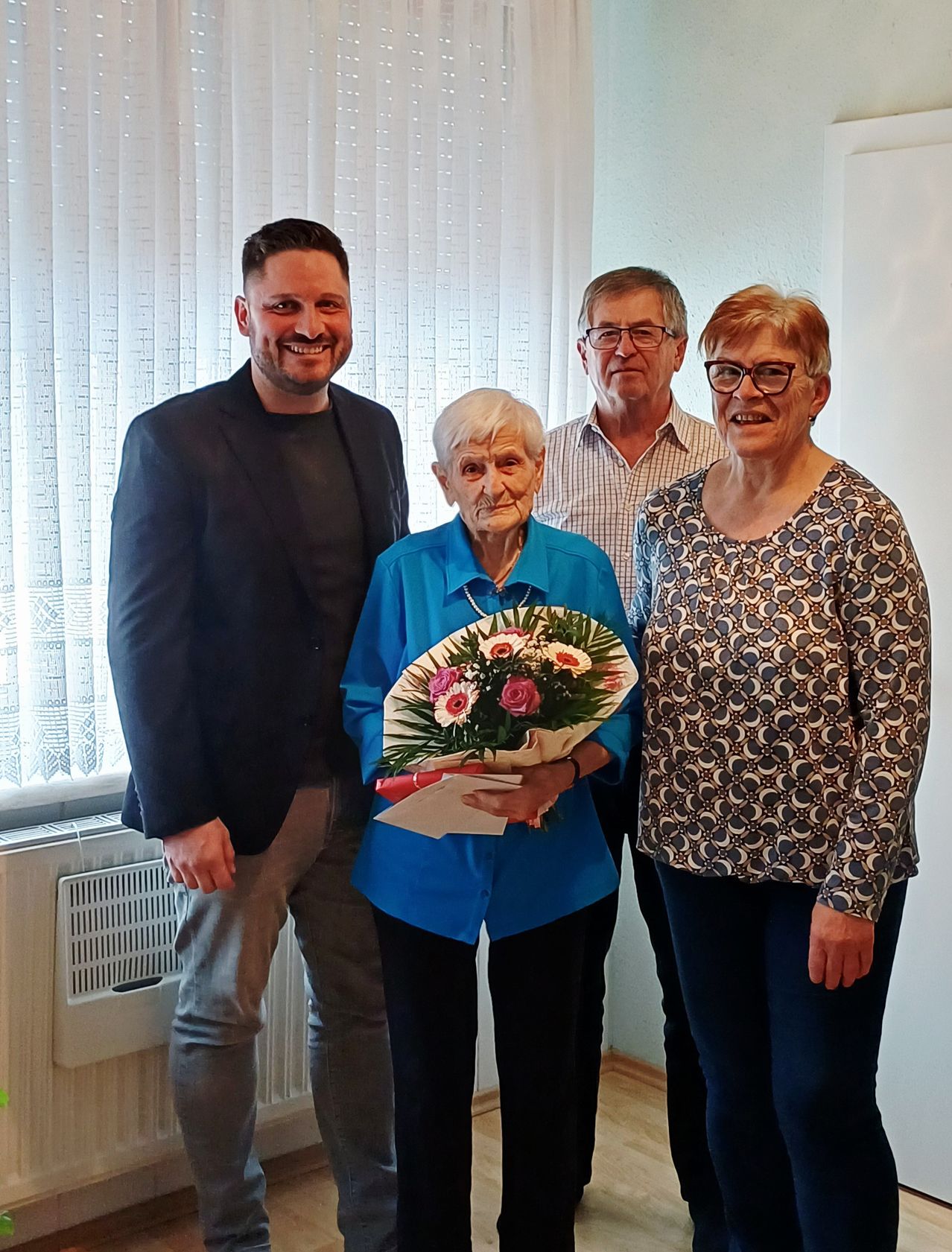 Vier Personen posieren für ein Foto in einem Raum. Eine ältere Frau hält Blumen und lächelt. Ein Mann und zwei Frauen stehen hinter ihr. Dahinter befindet sich ein Fenster mit weißen Vorhängen.