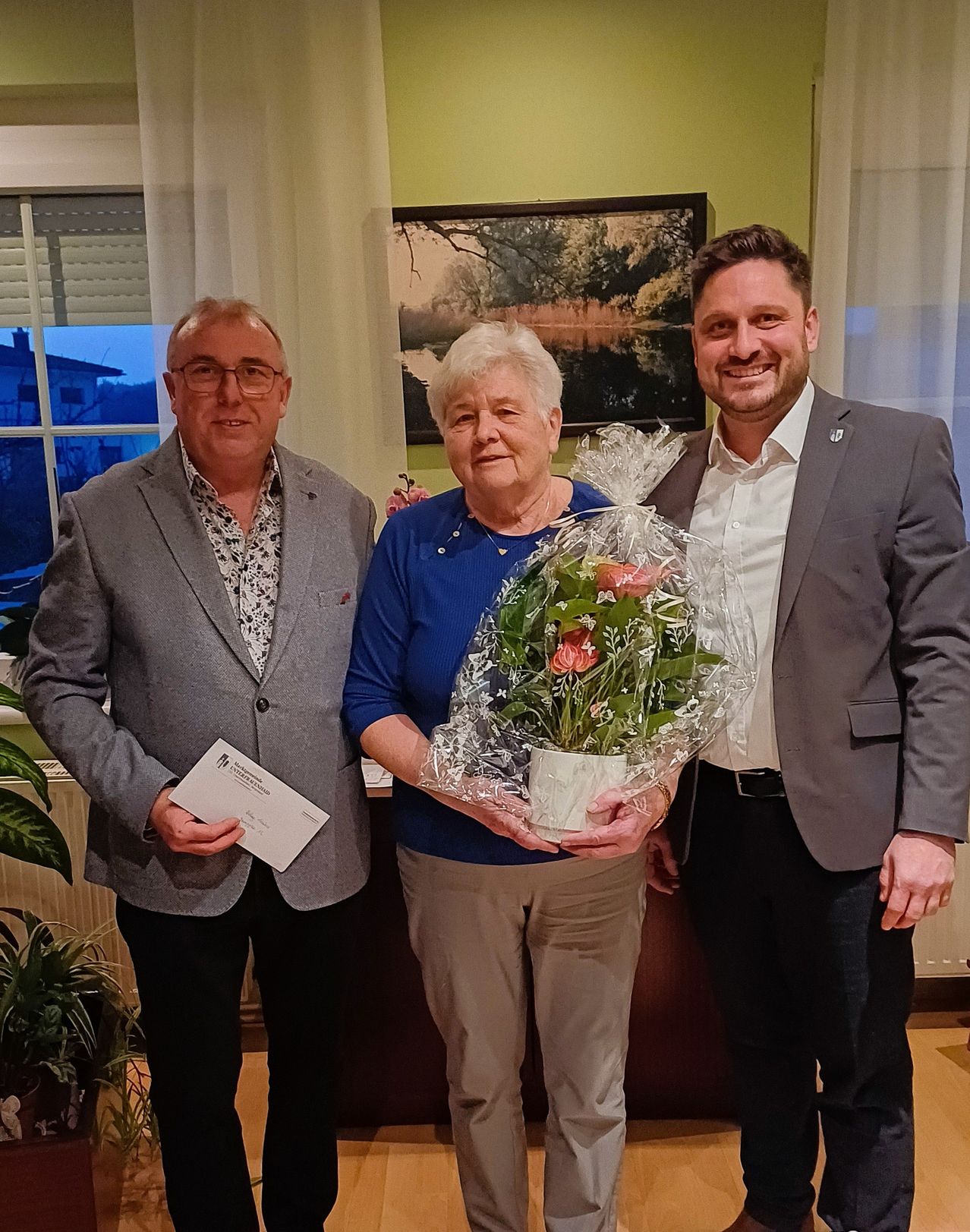 Drei Menschen stehen lächelnd im Innenraum, wobei eine Frau in der Mitte eine Blumenarrangement hält. Ein Mann auf der linken Seite hält ein Zertifikat. Hinter ihnen hängt ein Bilderrahmen an der Wand.