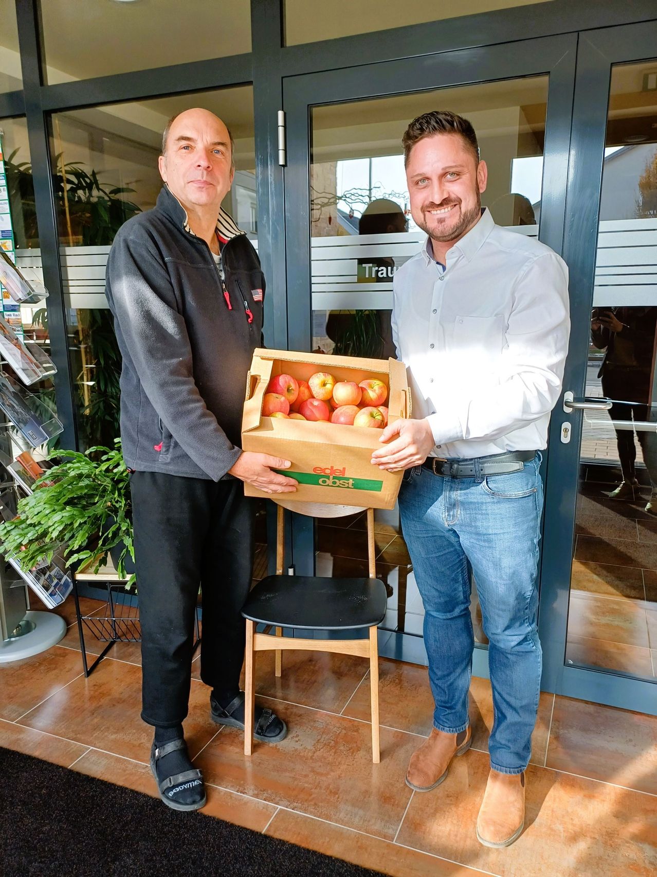 Zwei Männer stehen vor dem Eingang eines Geschäfts, einer hält eine Box mit Äpfeln, während der andere in die Kamera lächelt. Hinter ihnen befindet sich eine Glastür mit der Reflexion einer Person.