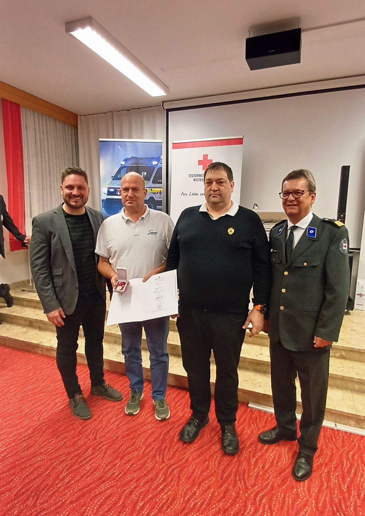 Vier Männer stehen auf einer Bühne mit rotem Teppich und halten ein Zertifikat. Dahinter befindet sich ein Banner mit einem Krankenwagen und einem Kreuz.