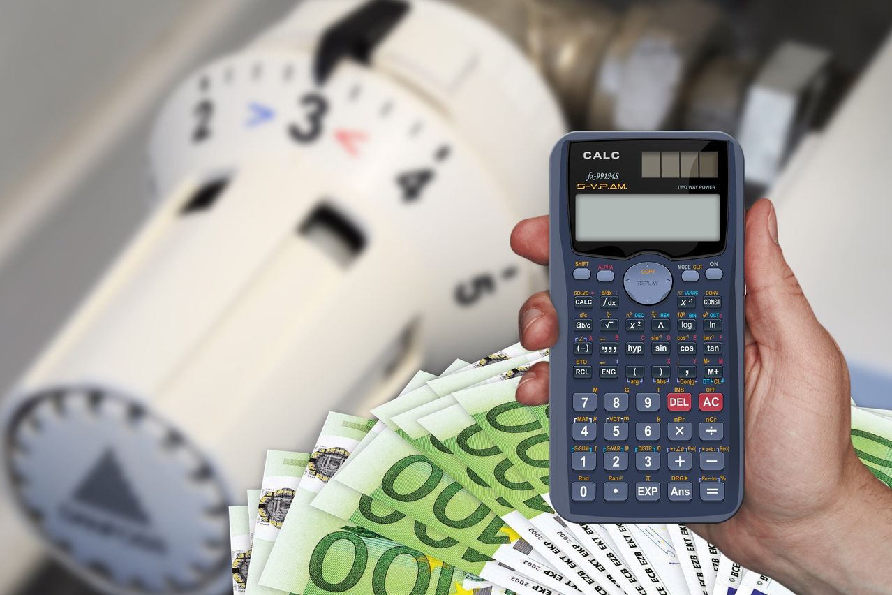 A person holds a calculator in front of a pile of cash and a clock with a blurred background. The calculator has a blue body and a screen with numbers and buttons.