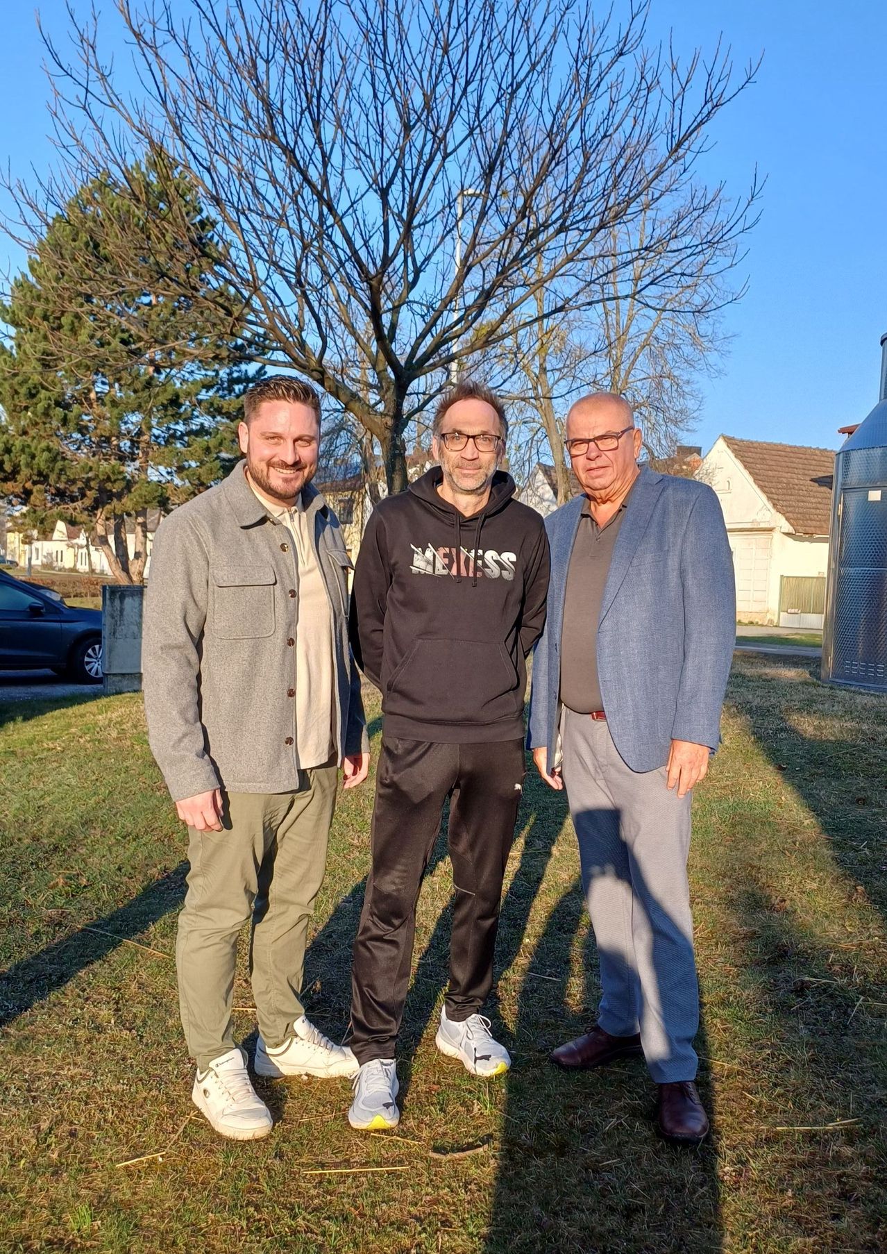 Three men are standing in a grassy area, smiling. Behind them is a house with a tree and a car.