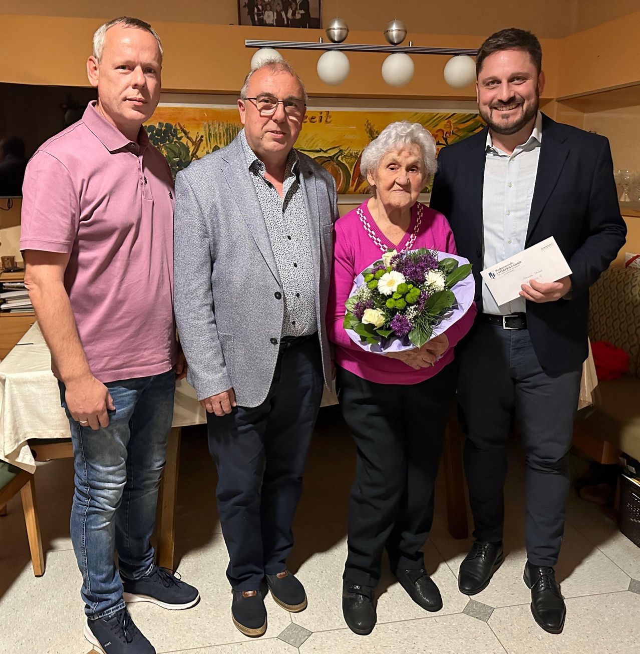 Four people are posing for a photo. An elderly woman holds a bouquet of flowers. The man on the right holds an envelope. The man on the left wears a pink shirt. The man in the middle wears a gray jacket.