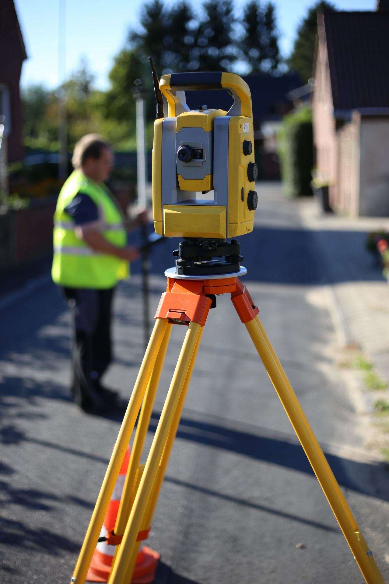 Ein Mann in einer neongrünen Weste steht auf der Straße und bedient ein Vermessungsgerät auf einem Stativ.