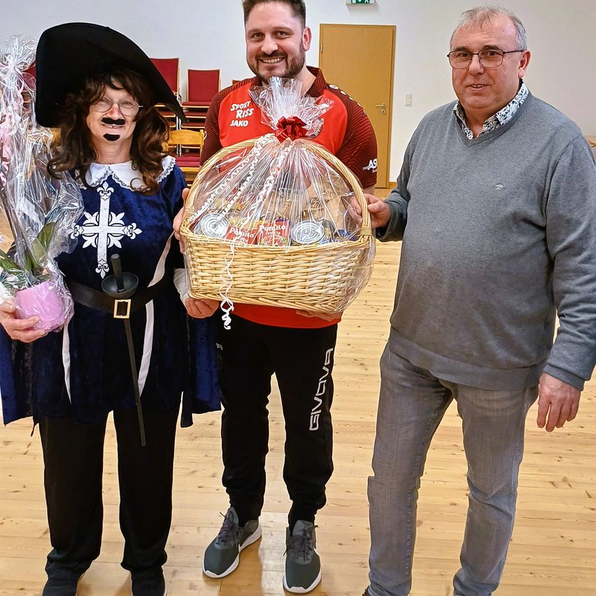 Three people stand in a room with wooden floors. A woman in a costume holds a bouquet. A man in sportswear holds a basket. Another man in a grey sweater stands next to them.