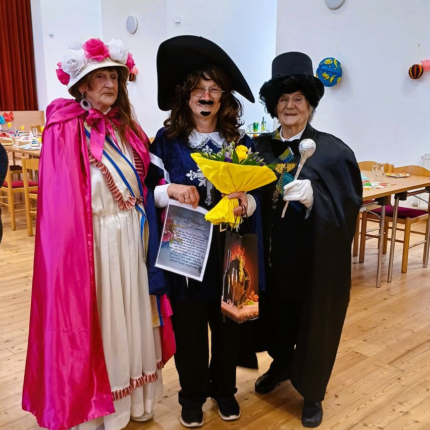 Three people dressed in costumes, one holding a bouquet of flowers and a gift bag, are standing together in a room. The room has wooden floors and tables.