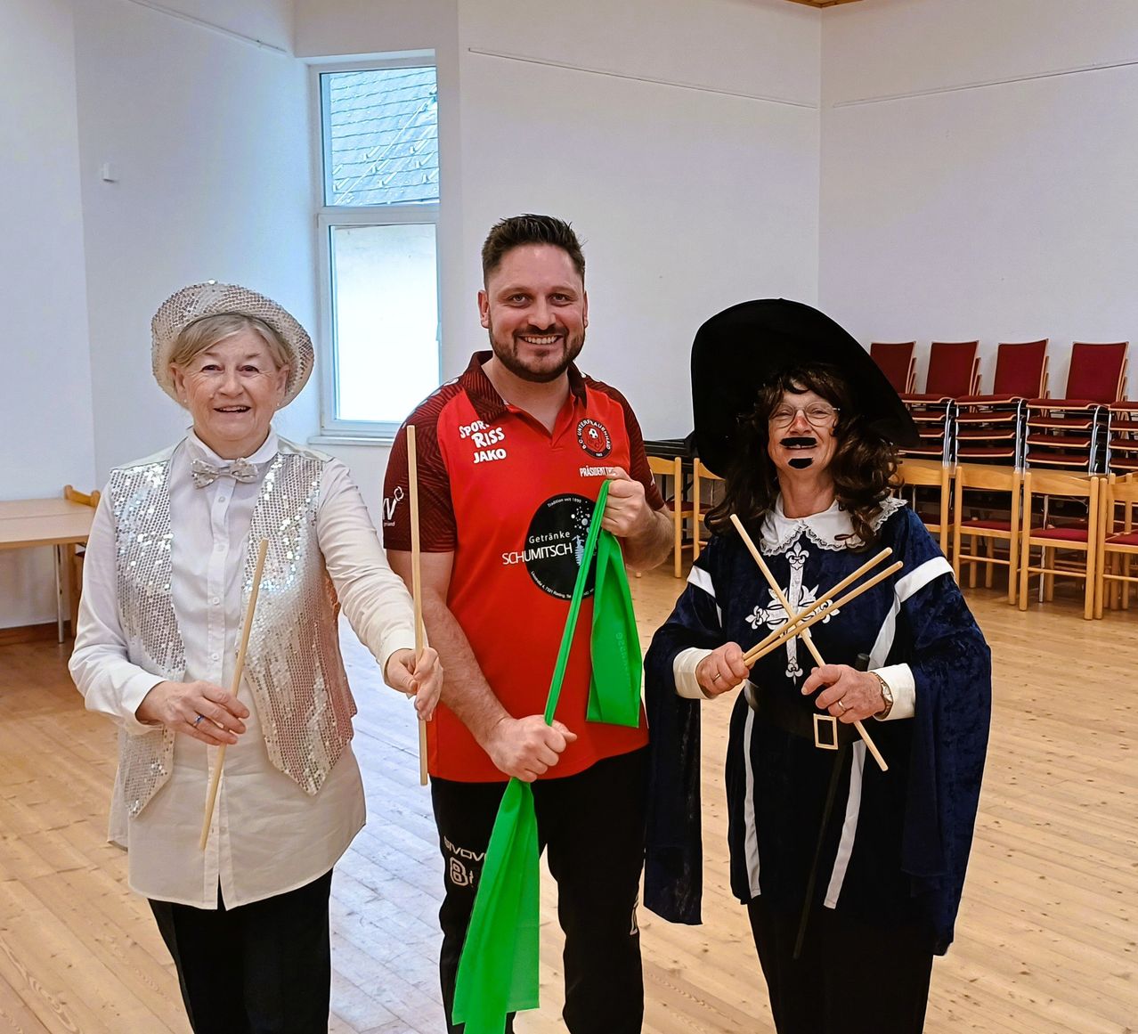 Three individuals stand in a room, holding sticks and smiling. The man in the middle wears a red shirt with a logo, and the women on either side wear hats and costumes. Behind them, chairs are arranged in rows.