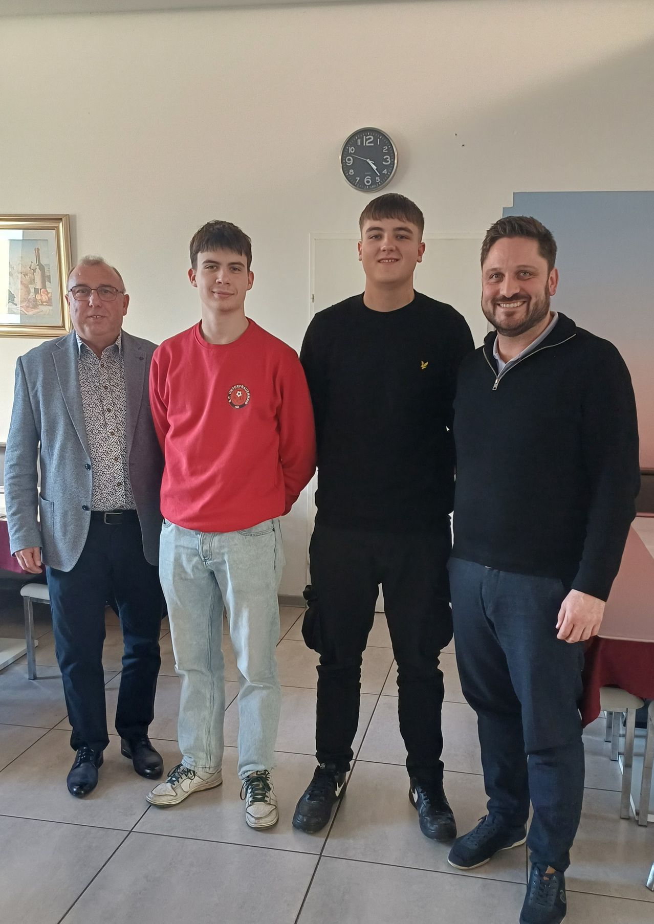 Vier Männer stehen in einem Raum zusammen. Die beiden in der Mitte lächeln, während die anderen nach vorne schauen. Dahinter befindet sich eine Uhr und ein Bilderrahmen.