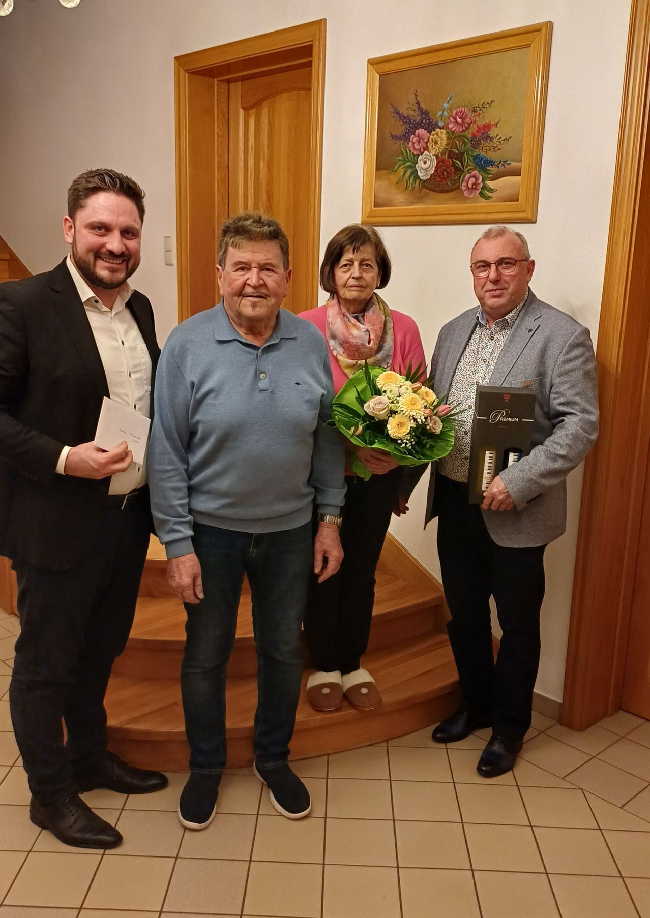Vier Personen stehen auf einer Treppe. Ein älterer Mann hält einen Blumenstrauß, und ein anderer hält ein Buch. Ein Mann auf der linken Seite hält ein Stück Papier. Hinter ihnen ist eine Holzwand mit einem Gemälde.