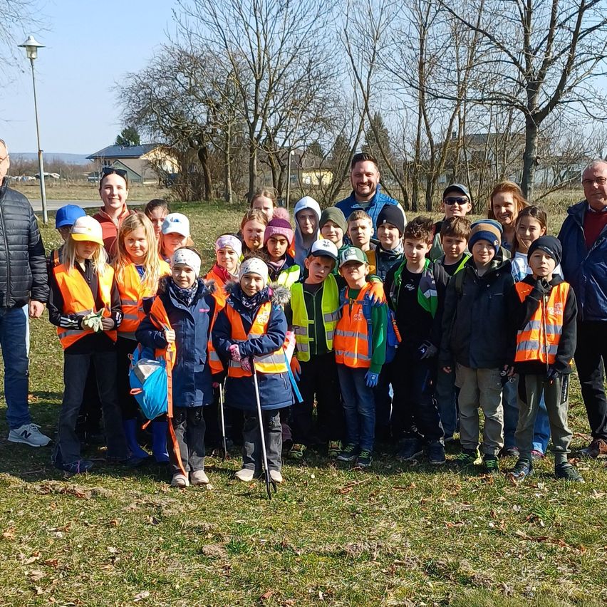 Eine Gruppe von Kindern und Erwachsenen in Warnwesten posiert für ein Foto auf einer Rasenfläche, mit Bäumen und Häusern im Hintergrund.