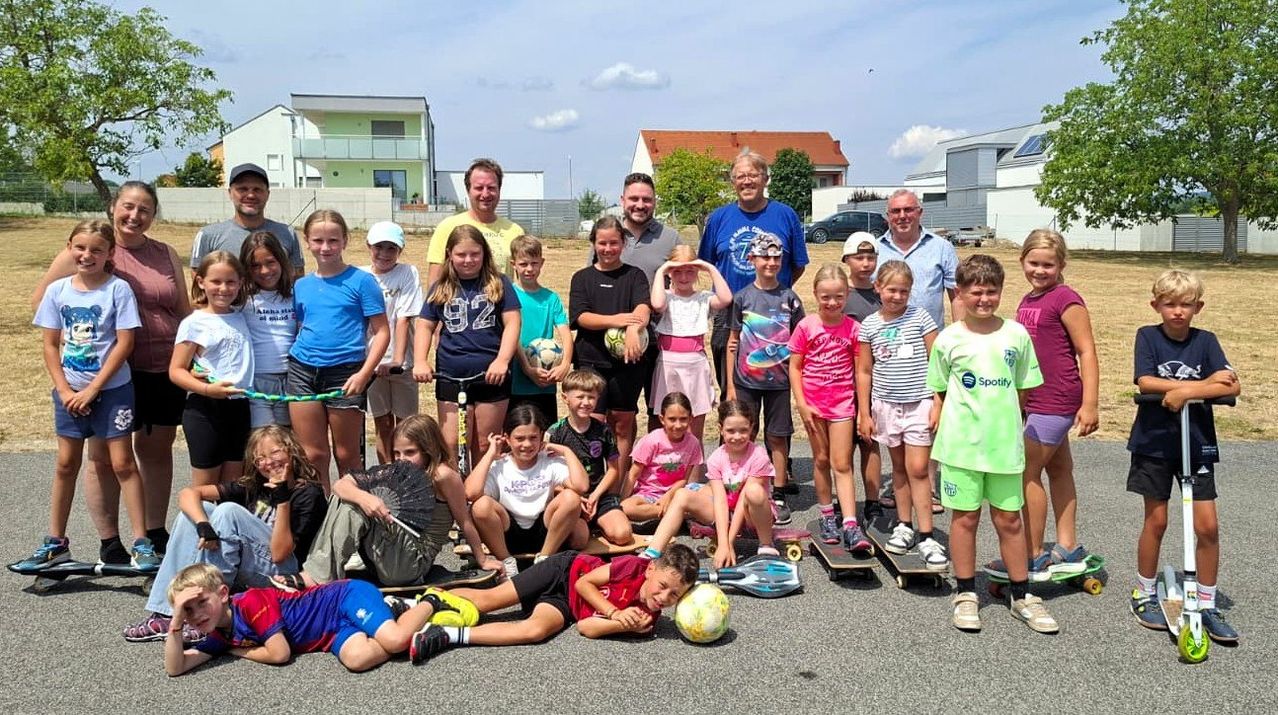 Eine Gruppe von Kindern und Erwachsenen posiert für ein Foto in einem Park, einige halten Sportgeräte und andere sitzen auf Skateboards.