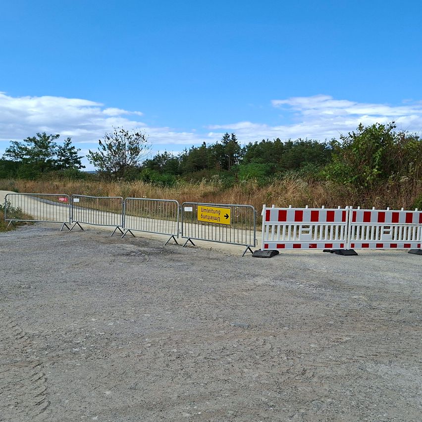 Eine Straße ist durch Metallbarrieren mit einem gelben Schild, das eine Umleitung anzeigt, blockiert. Das Gebiet ist von Bäumen und Gras umgeben unter einem klaren blauen Himmel.