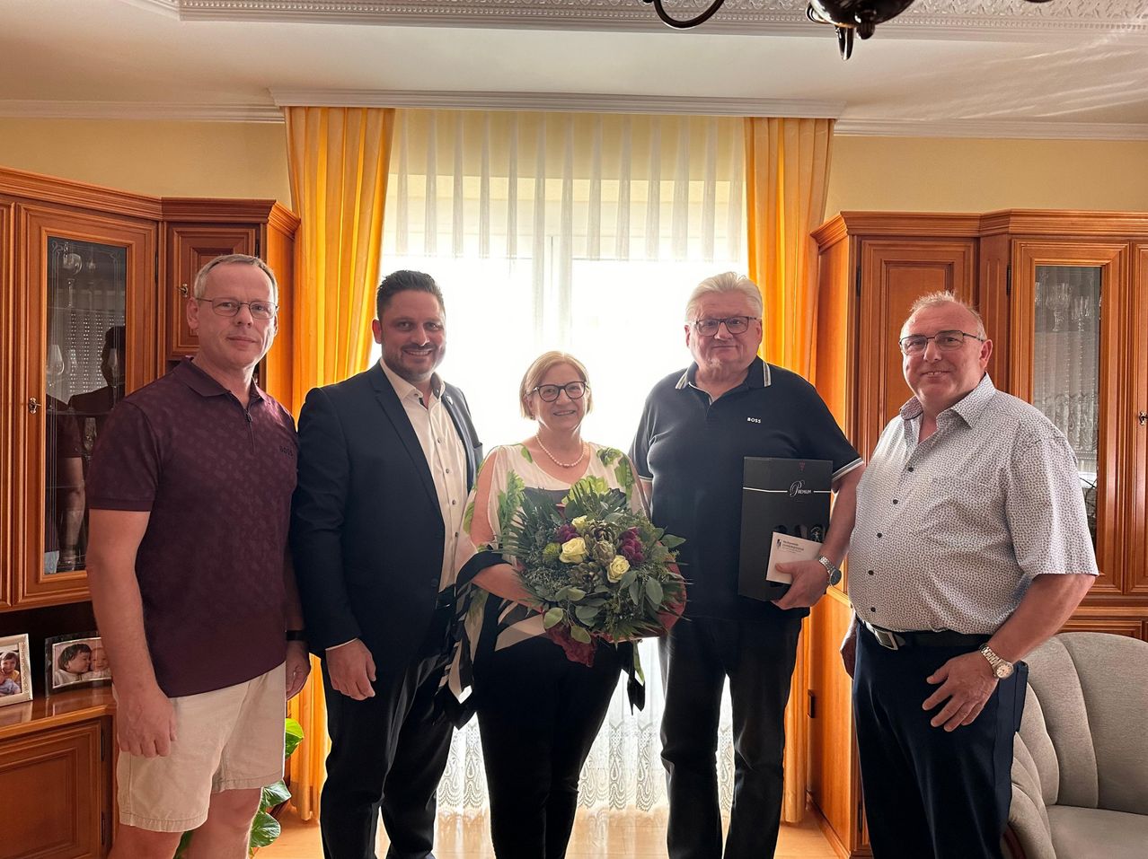 Fünf Männer und eine Frau stehen lächelnd für ein Foto in einem Raum. Die Frau hält einen Blumenstrauß. Hinter ihnen ist ein Fenster mit gelben Vorhängen.