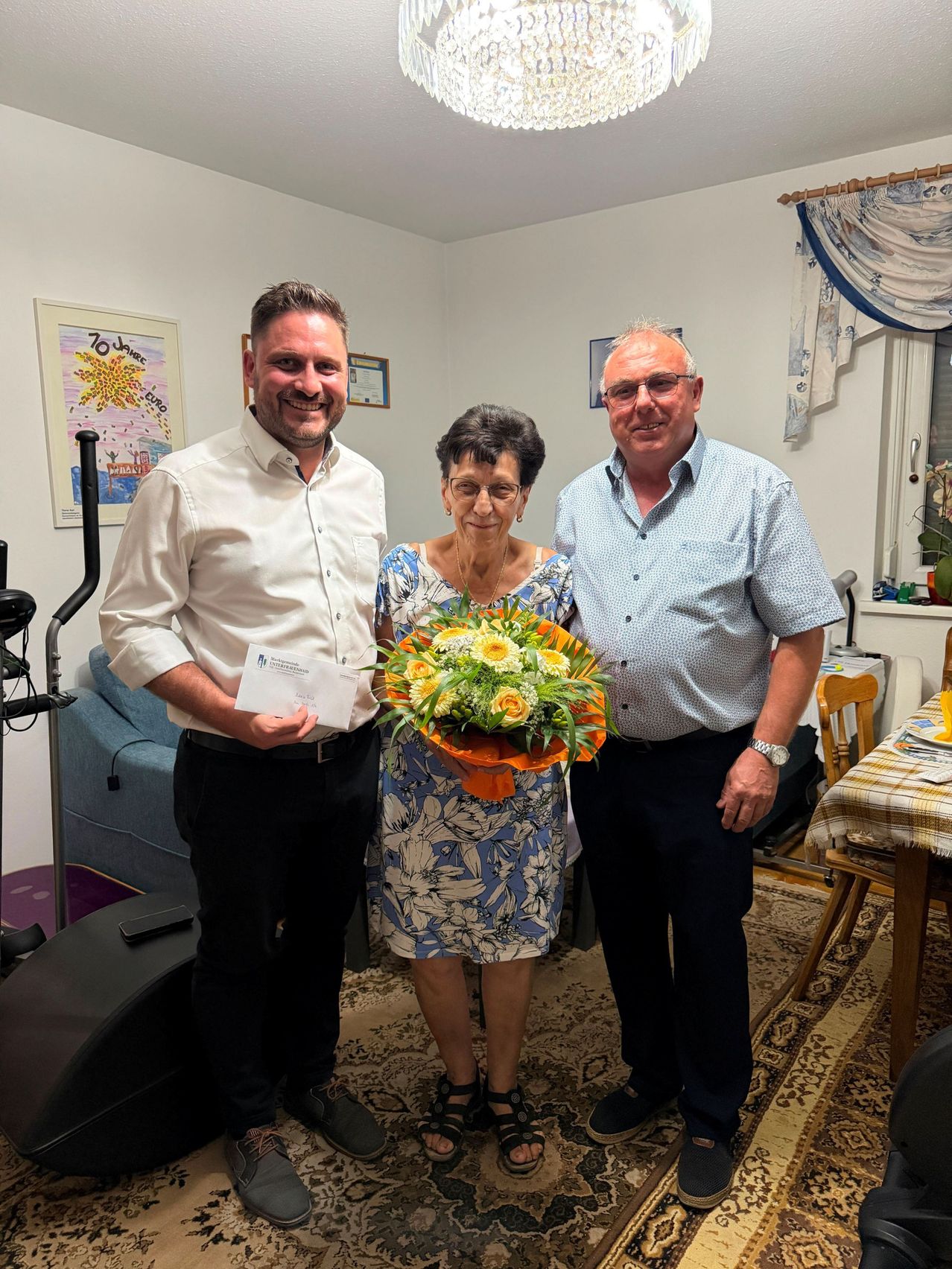 Drei Personen stehen in einem Raum und lächeln. Die Frau hält einen Blumenstrauß, während der Mann auf ihrer linken Seite ein Zertifikat hält. Der Mann auf ihrer rechten Seite trägt eine Brille. Hinter ihnen steht eine Couch, ein gerahmtes Bild und ein Tisch mit einer karierten Tischdecke.