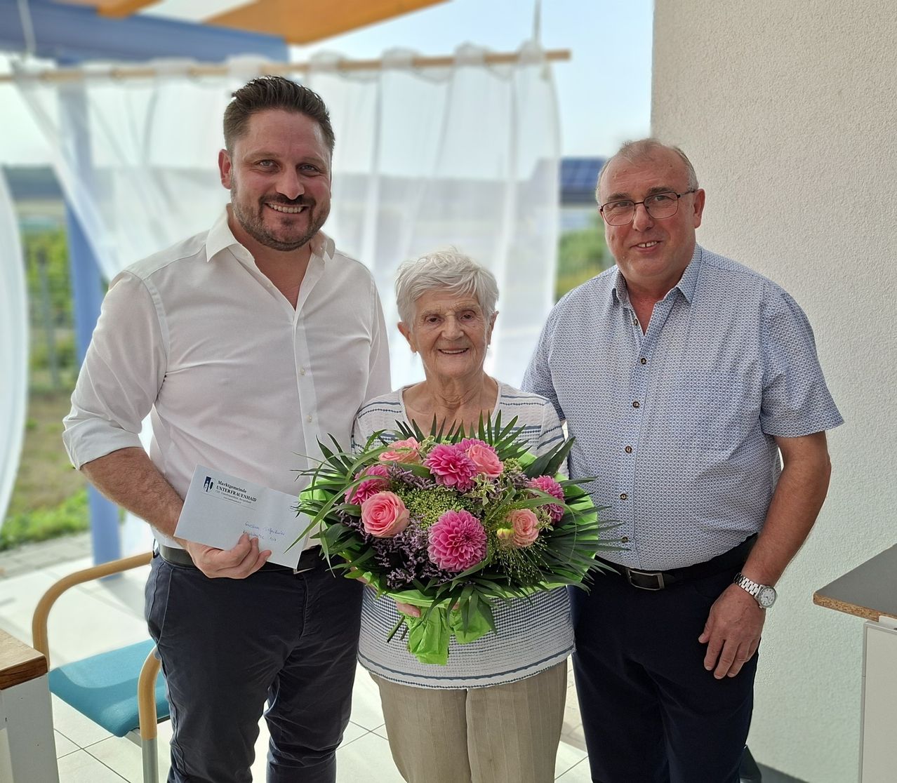 Drei Personen stehen lächelnd zusammen, eine Frau hält einen Blumenstrauß und ein Mann ein Zertifikat.