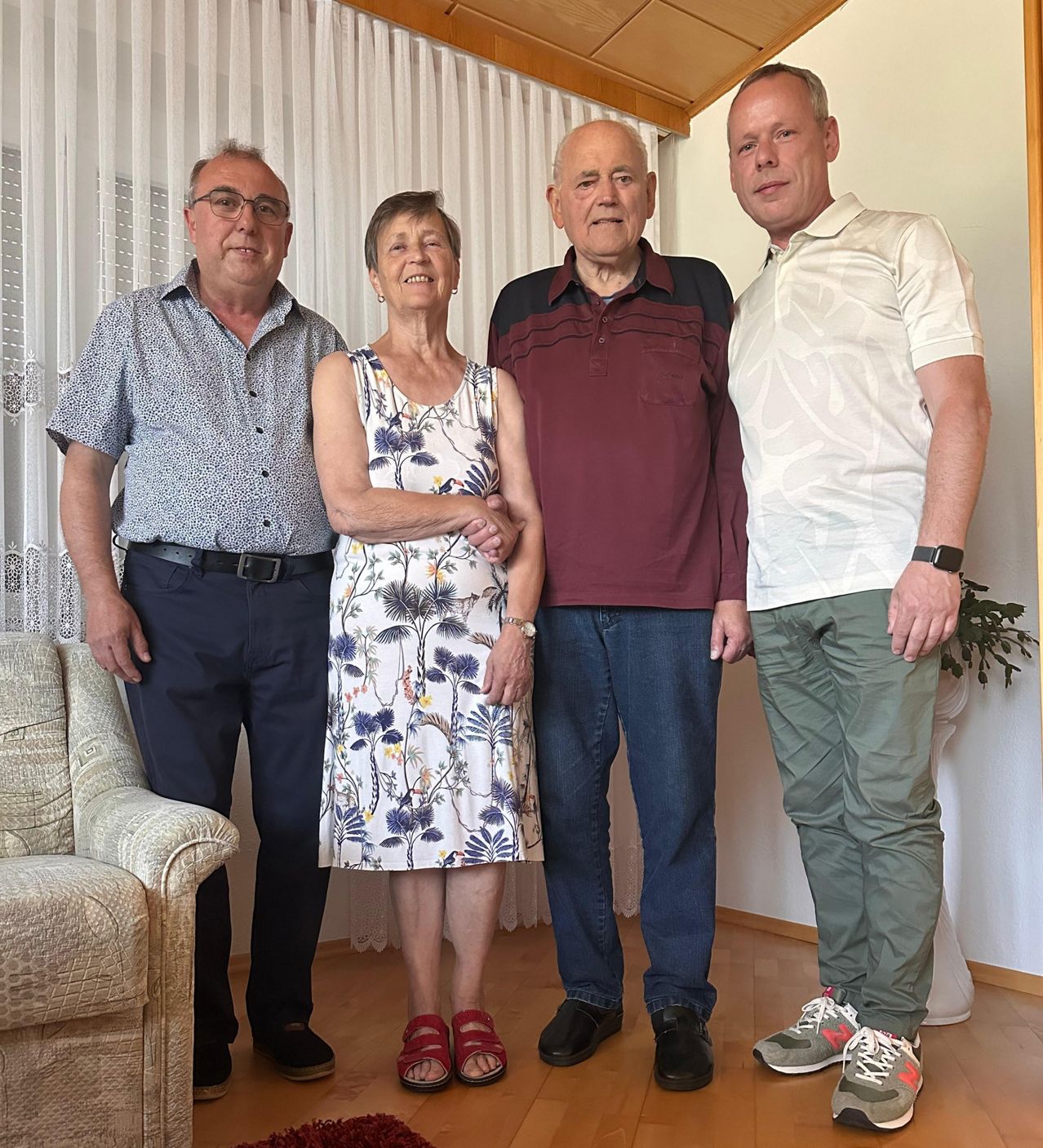 Vier Menschen stehen in einem Raum zusammen. Eine Frau in einem Blumenkleid lächelt, während die Männer lässig gekleidet sind. Ein Mann trägt eine Brille, ein anderer eine Uhr und der dritte ein Armband. Der Boden ist aus Holz und ein Sofa und eine Pflanze sind im Hintergrund zu sehen.