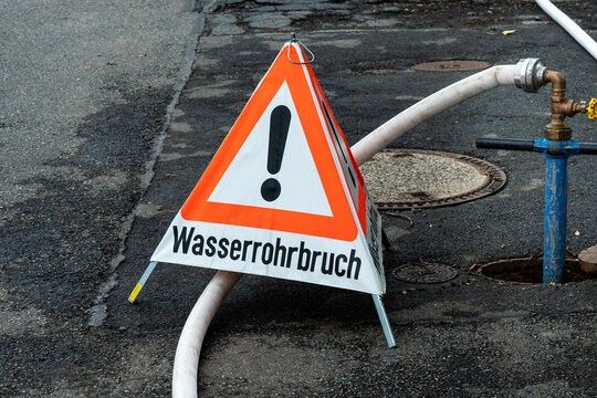 Eine beschädigte Wasserleitung mit einem Warnschild auf der Straße. Die Leitung ist mit einem Wasserhahn verbunden.