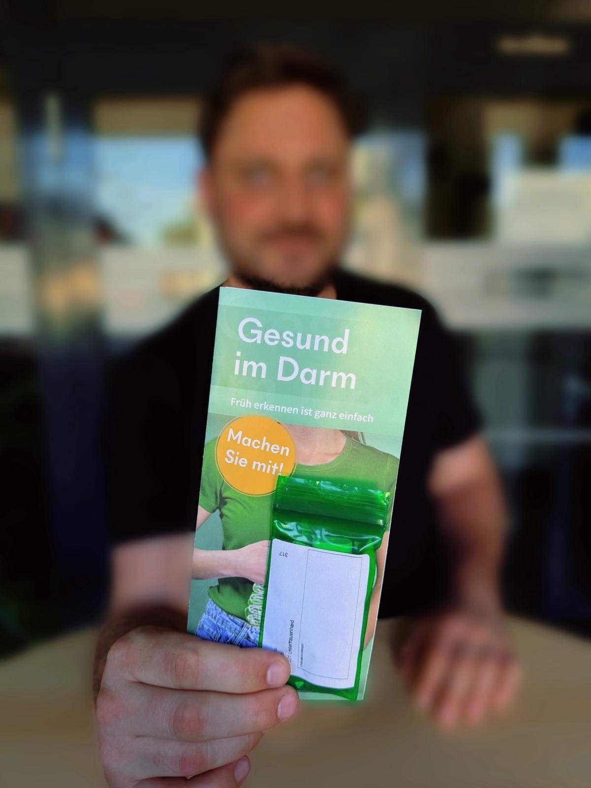 Ein Mann hält eine Broschüre mit dem Titel 'Gesund im Darm' und einem angehängten Plastikbeutel vor einem unscharfen Hintergrund.