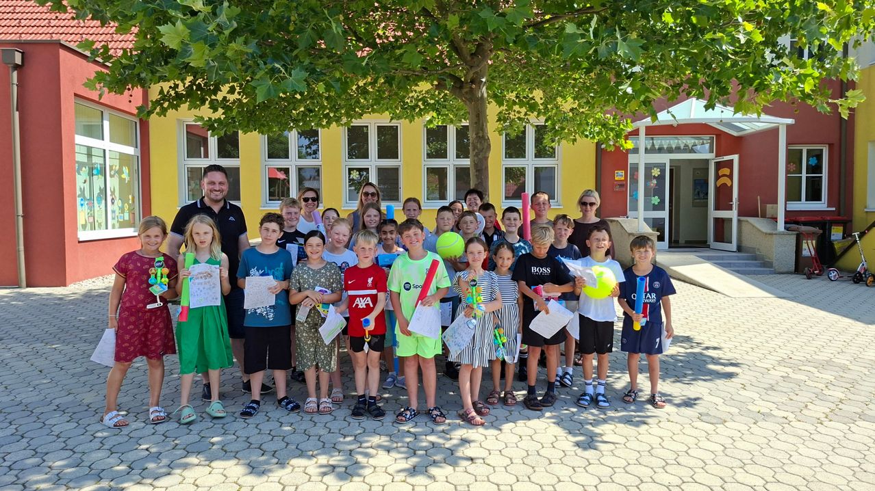 A group of children and adults stand under a tree outside a yellow building, holding papers and sports equipment. Some wear sunglasses, and the adults smile.