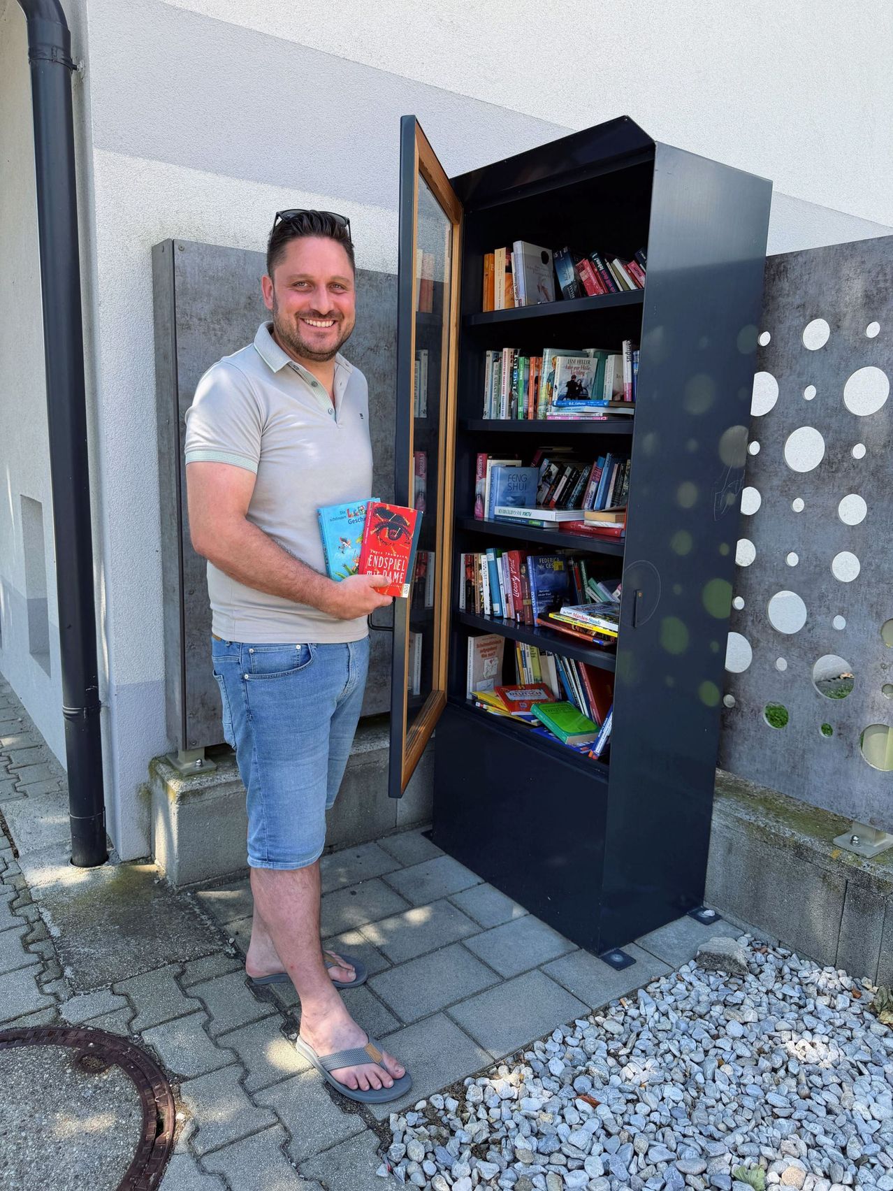 A man holds two books in front of a bookshelf. He stands outside with a smiling face. The bookshelf is filled with many books.