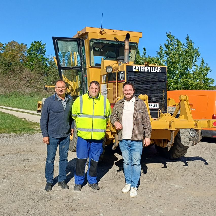 Drei Männer stehen vor einem gelben Caterpillar-Grader auf einem Feldweg
