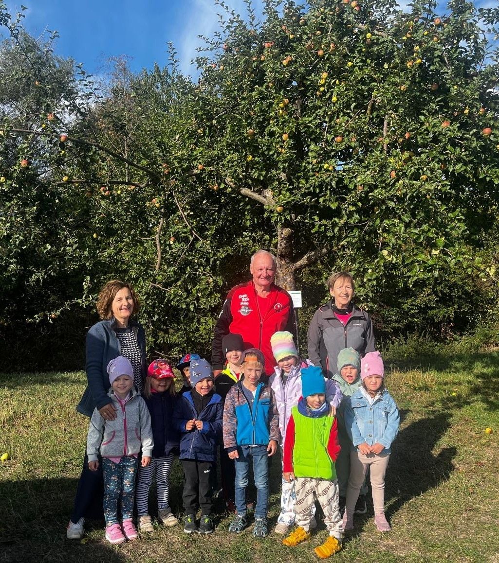 Eine Gruppe von Kindern und Erwachsenen steht vor einem Apfelbaum und lächelt für ein Foto.