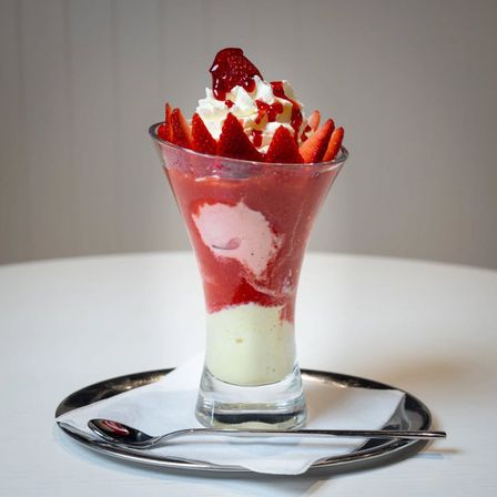 Ein hoher Glasbecher gefüllt mit Erdbeereis und Schlagsahne, garniert mit Erdbeerscheiben, auf einem Teller mit einem Löffel.