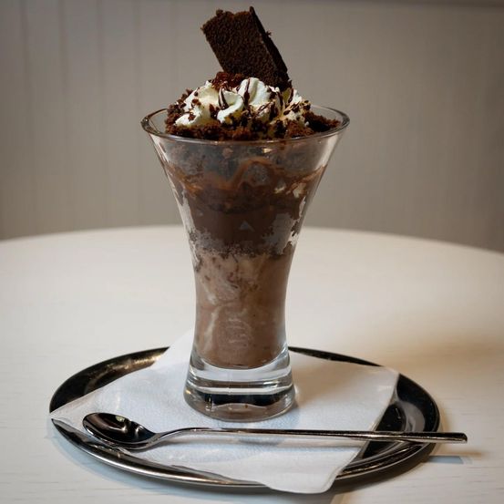 Ein Schokoladeneisbecher mit Schlagsahne und Schokoladenraspeln, garniert mit einem Brownie auf der Oberseite, serviert in einem hohen Glas auf einem weißen Teller mit einem Löffel.