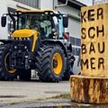 Ein gelber Traktor mit großen Reifen steht in der Nähe eines Gebäudes. Ein hölzerner Pfosten mit geschnitzten Buchstaben steht in der Nähe. Menschen gehen hinter dem Traktor.