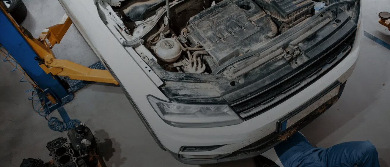 Eine Nahaufnahme der Fahrzeugfront mit offenem Motorhauben, der Motor ist zu sehen. Eine Person in blauer Kleidung untersucht den Motor.