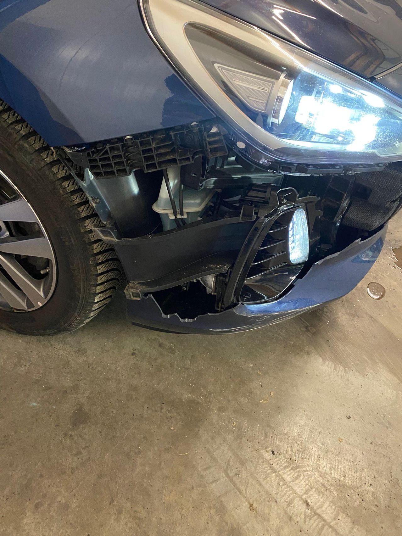 Nahaufnahme des beschädigten Stoßfängers eines blauen Autos, das den Scheinwerfer, den Kühlergrill und einen Teil des Motorraums zeigt.