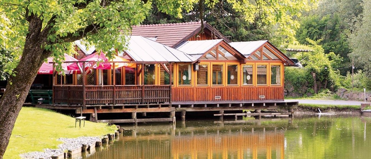 Ein Holzhaus mit braunem Dach steht am Seeufer. Das Haus hat Glasfenster und rote Sonnenschirme. Die Reflexion des Hauses ist im Wasser.