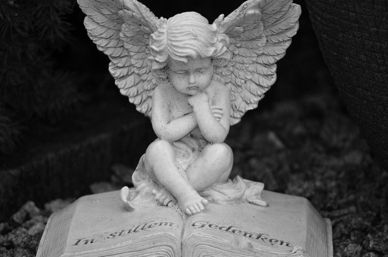 Eine kleine Statue eines Cherubs sitzt auf einem offenen Buch. Die Statue ist weiß und das Buch ist grau. Der Cherub betet mit gefalteten Händen.