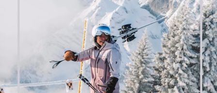 Ein Skifahrer trägt Skier und Stöcke und steht auf einem verschneiten Berg, lächelt und posiert mit verschneiten Bergen im Hintergrund.
