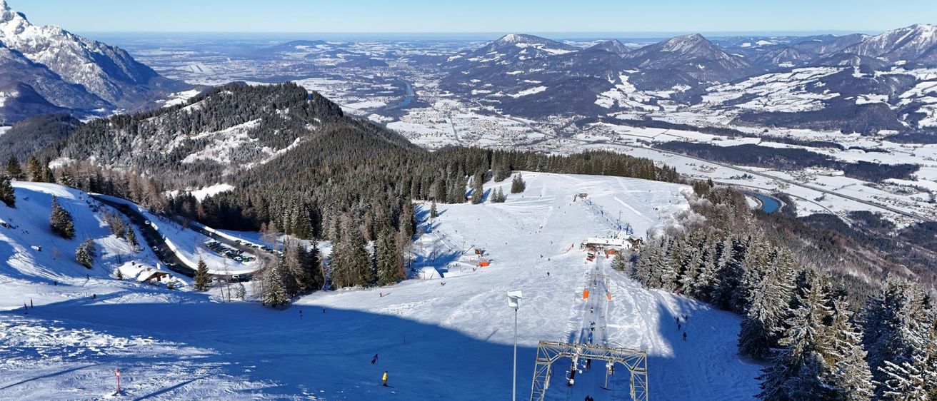 Luftaufnahme eines verschneiten Skigebiets mit Skifahrern auf den Pisten, umgeben von Bäumen und Bergen. Im Vordergrund befindet sich eine Skiliftanlage.