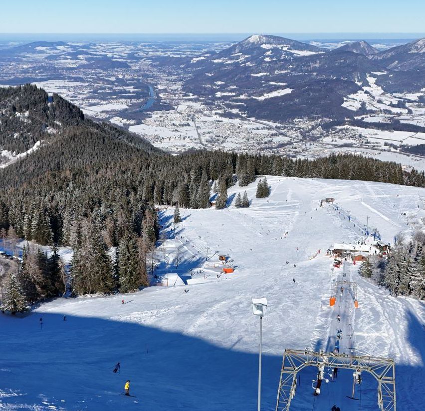 Luftaufnahme eines verschneiten Skigebiets mit Skifahrern auf den Pisten, umgeben von Bäumen und Bergen. Im Vordergrund befindet sich eine Skiliftanlage.