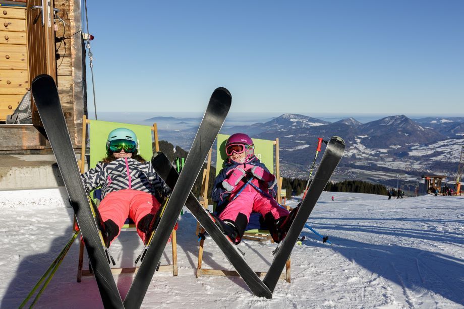Zwei Kinder ruhen in grünen Stühlen mit ihren Skiern auf einem verschneiten Berg und genießen die Aussicht auf entfernte Berge.