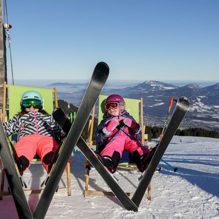 Zwei Kinder ruhen in grünen Stühlen mit ihren Skiern auf einem verschneiten Berg und genießen die Aussicht auf entfernte Berge.